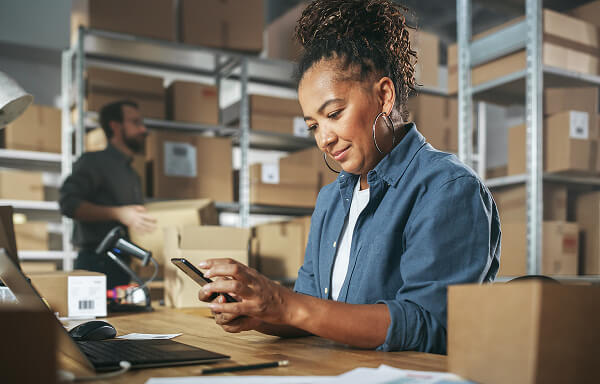  Woman in warehouse looking at mobile device with laptop open on the desk