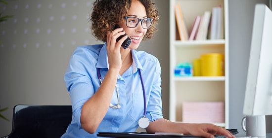 Doctor sitting at desk while talking on the phone and looking at her desktop device