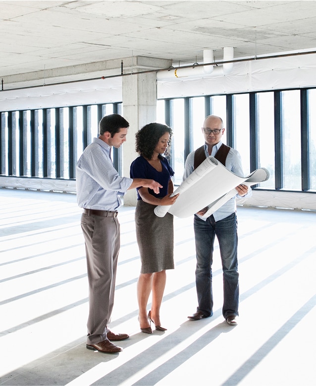 Three colleagues in an empty building holding large blueprints.