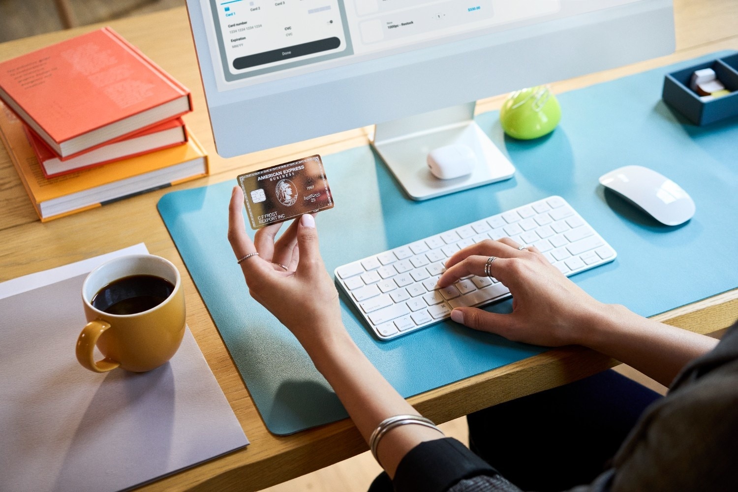 A person holds an American Express Business Platinum Card in one hand while using a computer keyboard at a desk with books and coffee.