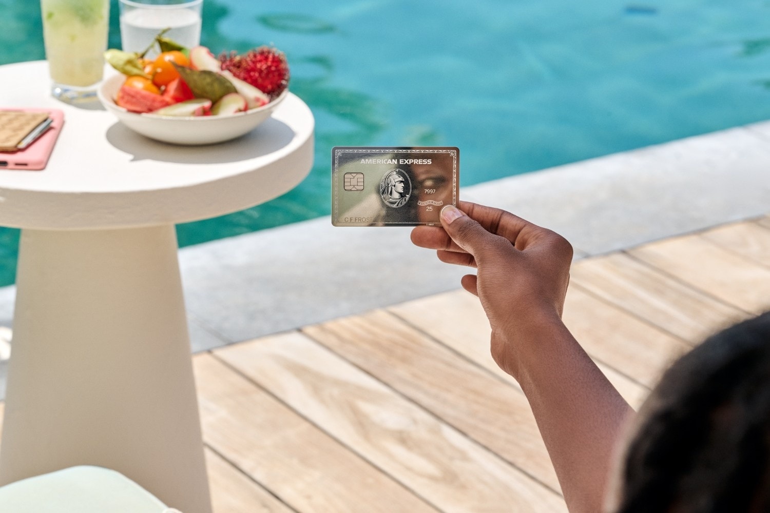 A person holds an American Express Platinum Card near a pool, next to a table with fruit, water, and drinks.