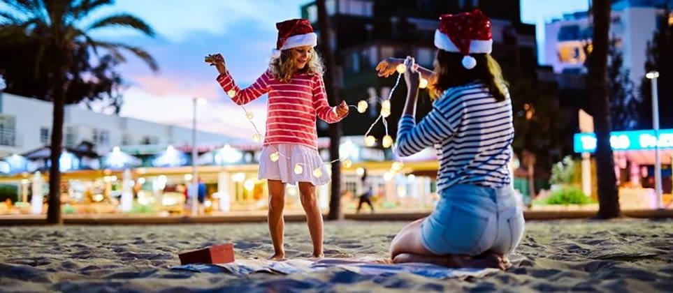 Woman and child wearing Santa hats, playing with Christmas lights on a tropical beach