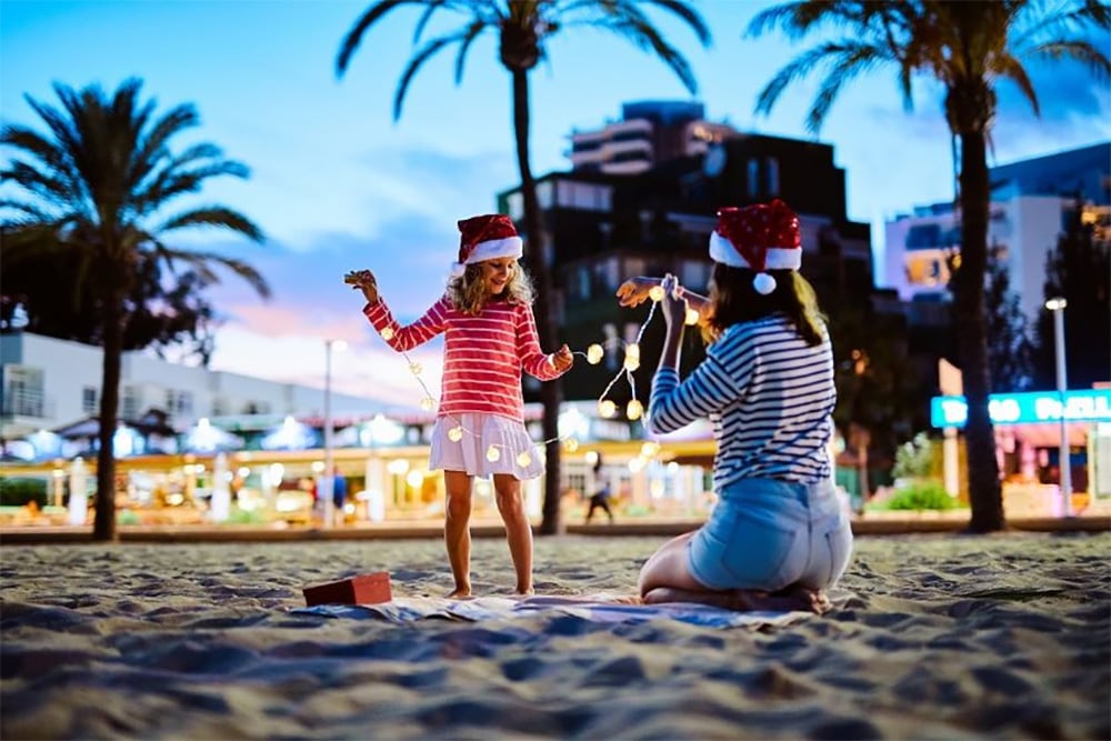 Woman and child wearing Santa hats, playing with Christmas lights on a tropical beach