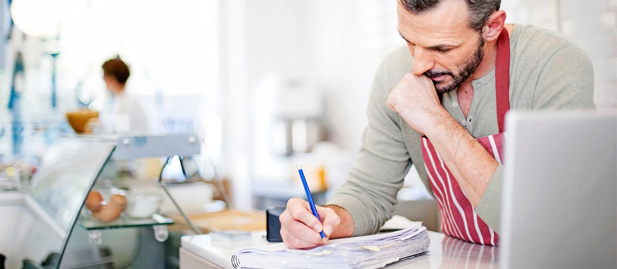 Man in an apron is writing in the journal inside a grocery store behind the register table