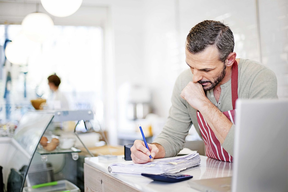 Man in an apron is writing in the journal inside a grocery store behind the register table