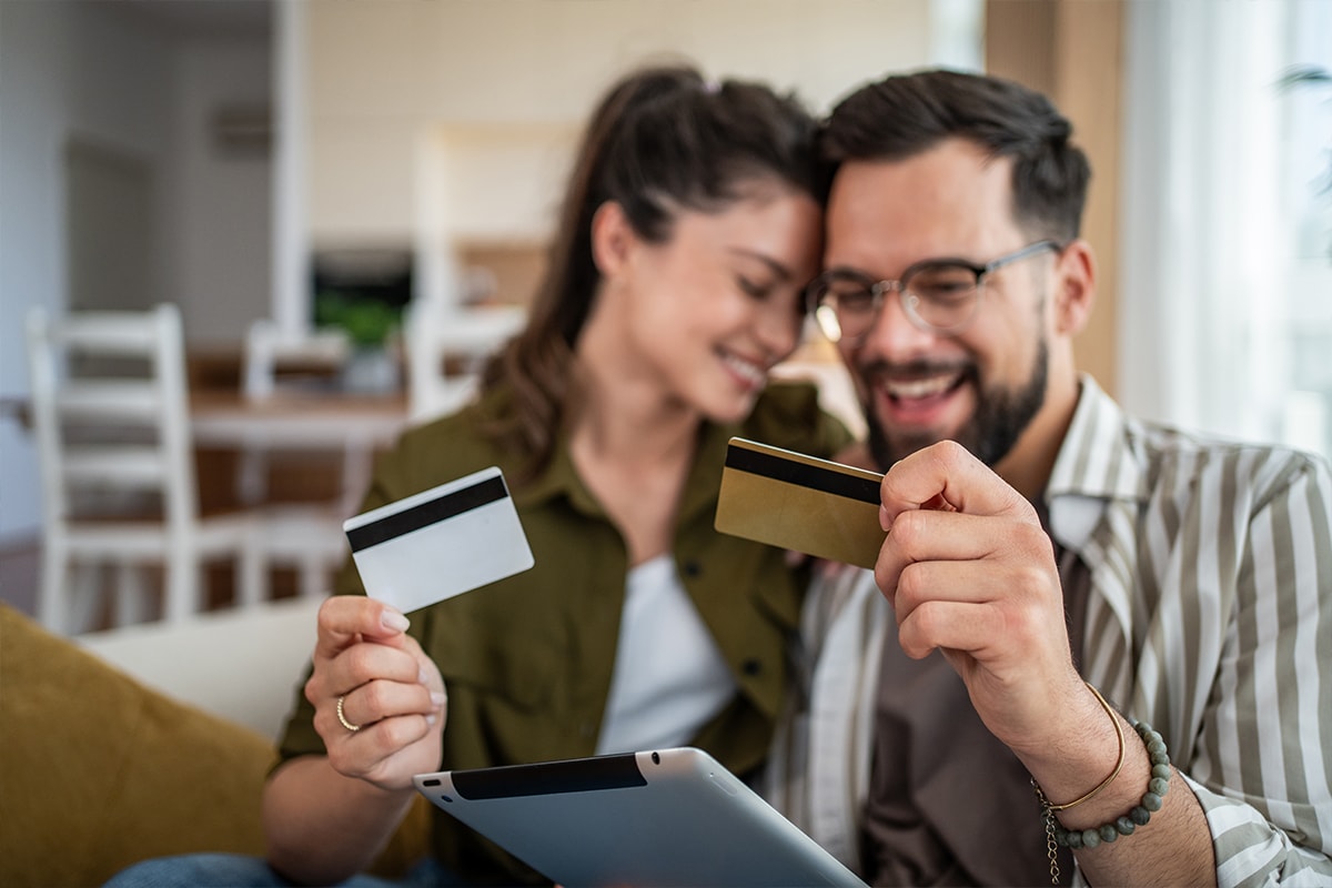 A person in their mid-thirties sitting at an island kitchen countertop with a laptop, smiling at the two credit cards held in each hand.