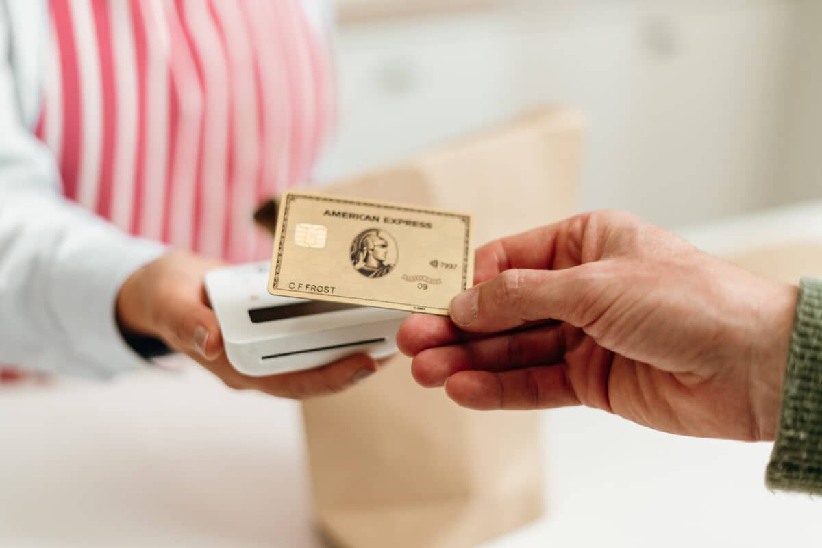 A person handing an Amex Gold card to a cashier using a contactless card reader at a store checkout.