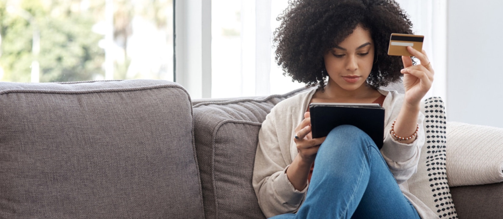Woman sitting on her couch looking at her tablet holding a credit card