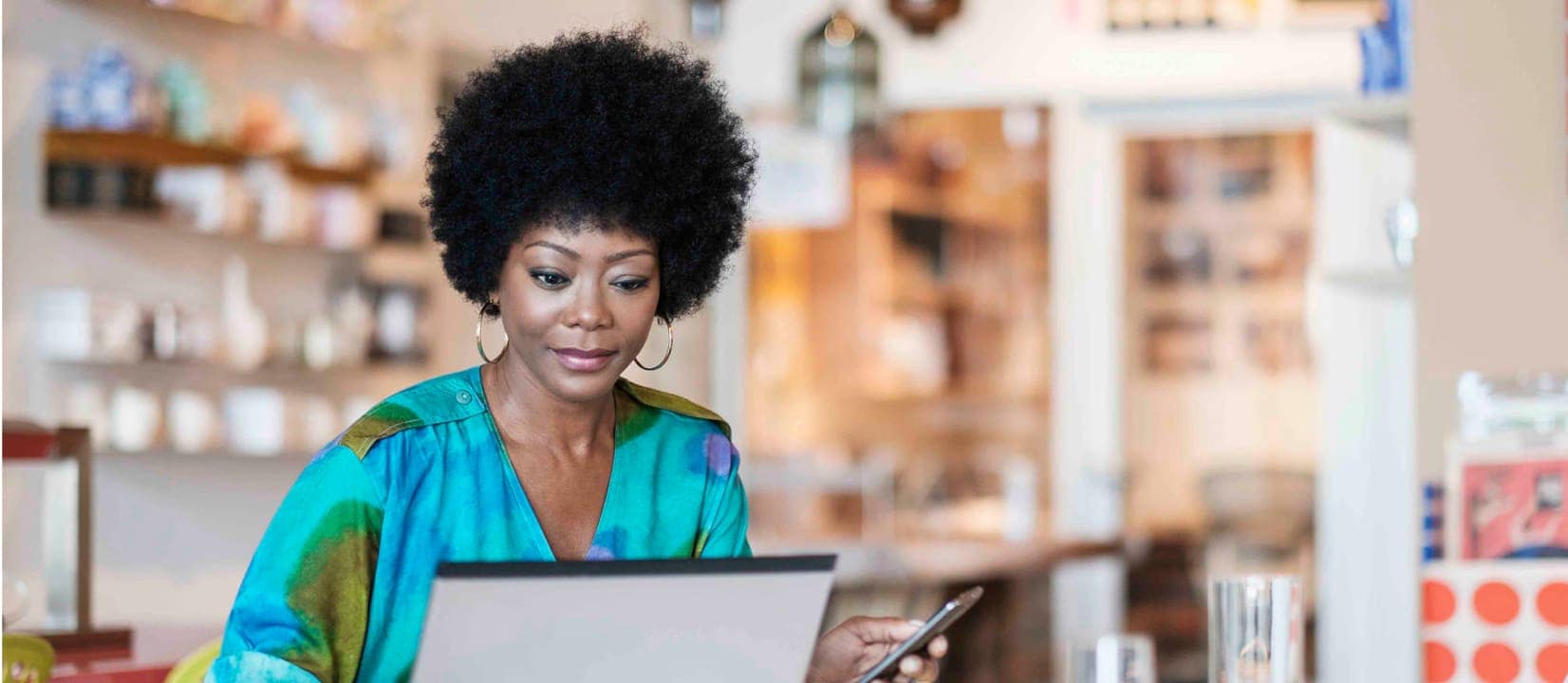 While sitting in a shop a woman uses her laptop and phone to make a car payment