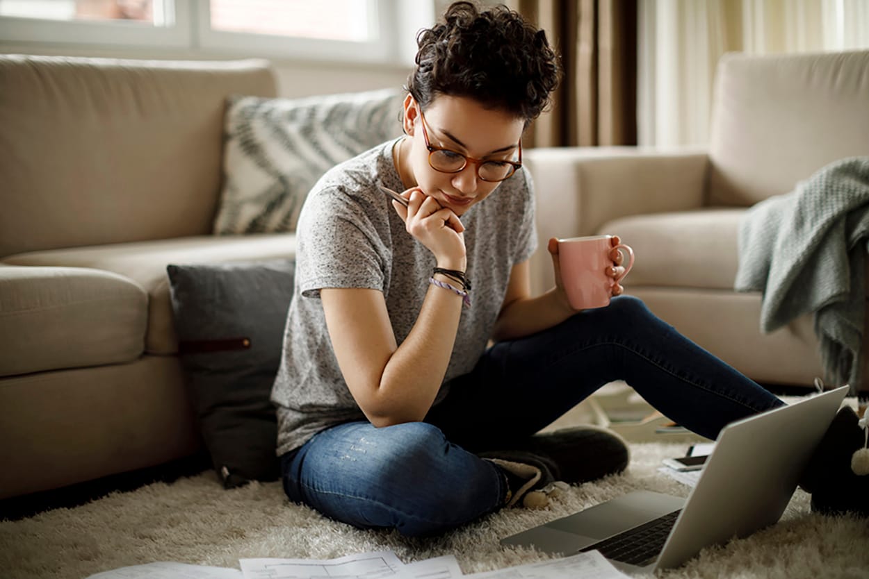 A woman sitting on the floor of her living room looking over bills with her laptop in front of her