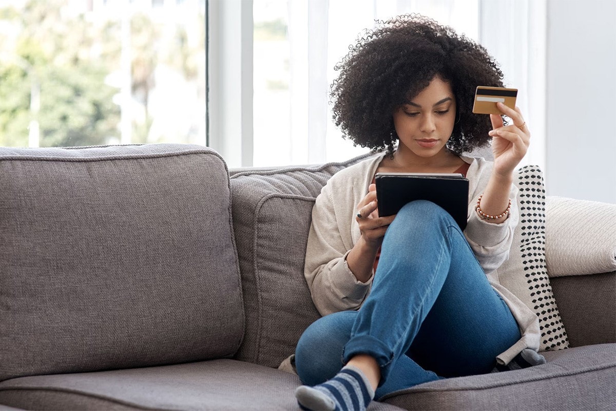 Woman sitting on her couch looking at her tablet holding a credit card