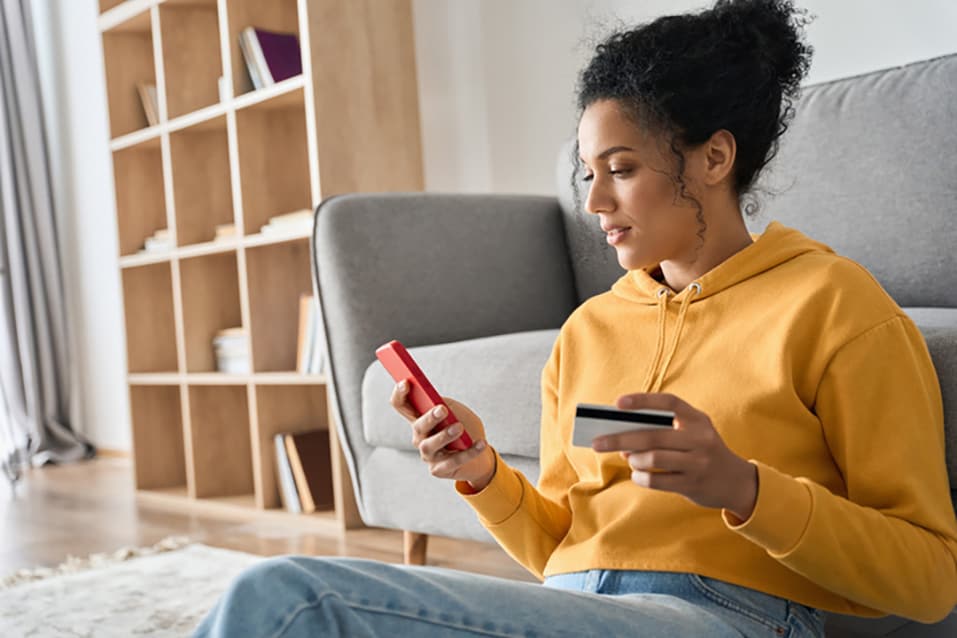 Woman in a yellow hoodie sitting on the floor and looking at a cellphone while holding a credit card