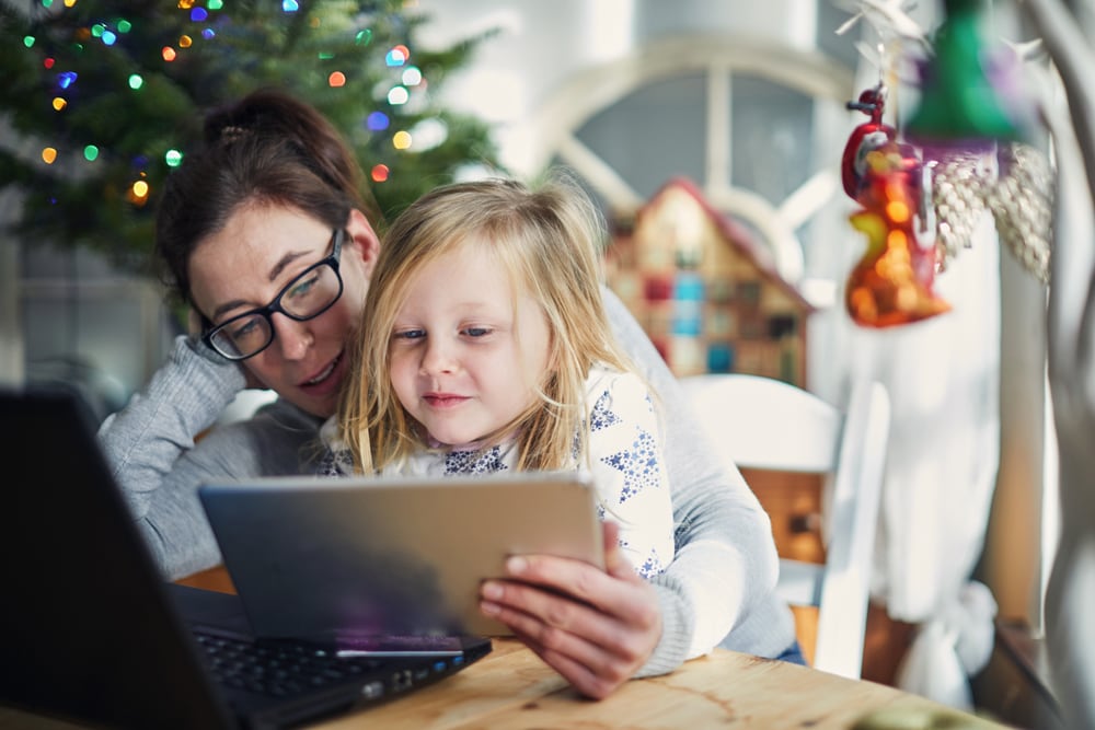 A mother holding her daughter while they both look at a tablet in front of Christmas tree