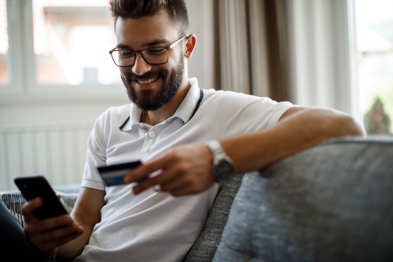 Man smiling and sitting on his couch holding his credit card while looking at his phone