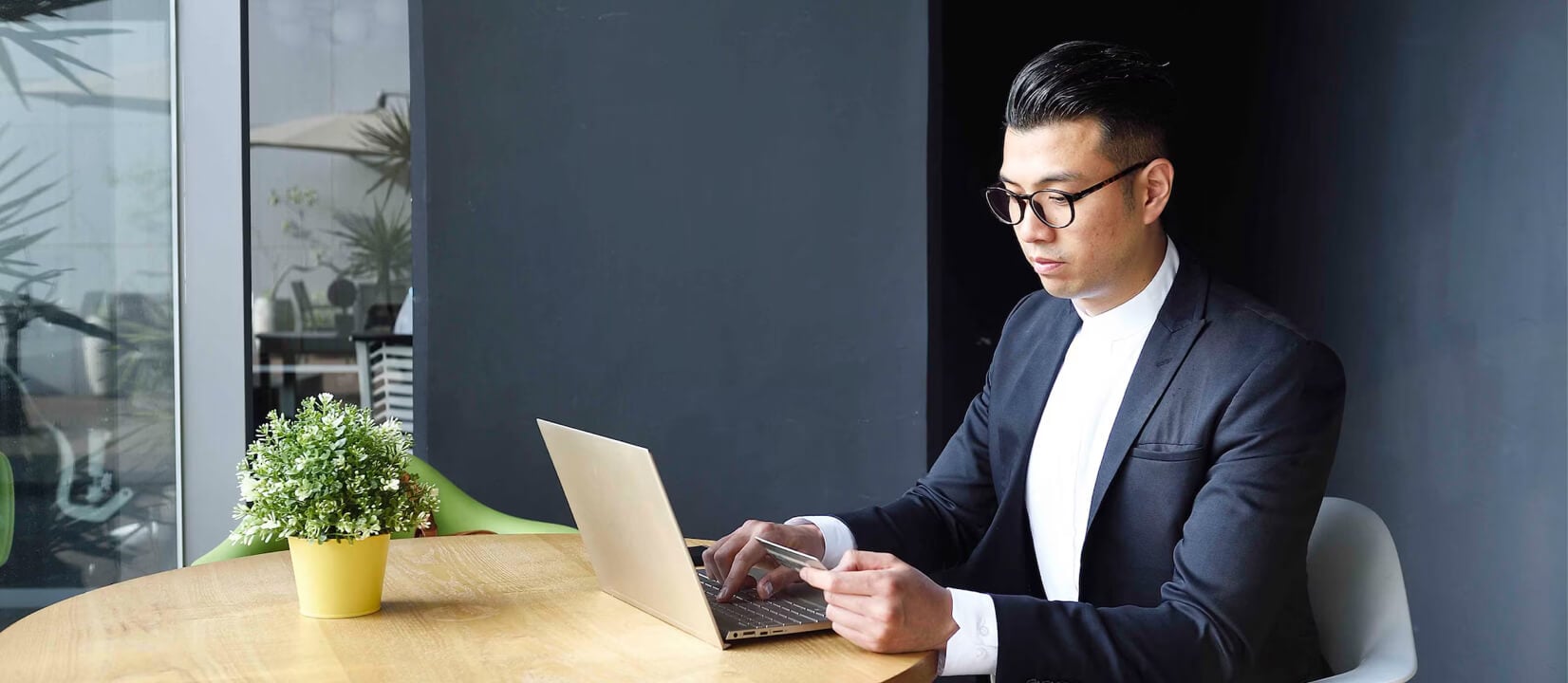 A man sits outside at a table while using his laptop and looking at his credit card.