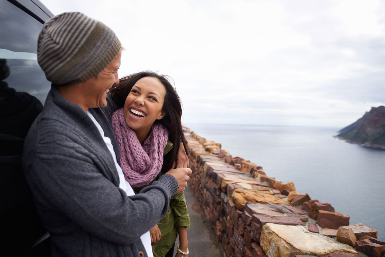A smiling man and woman stand together in front of a car, highlighting the advantages of credit card rental car insurance.