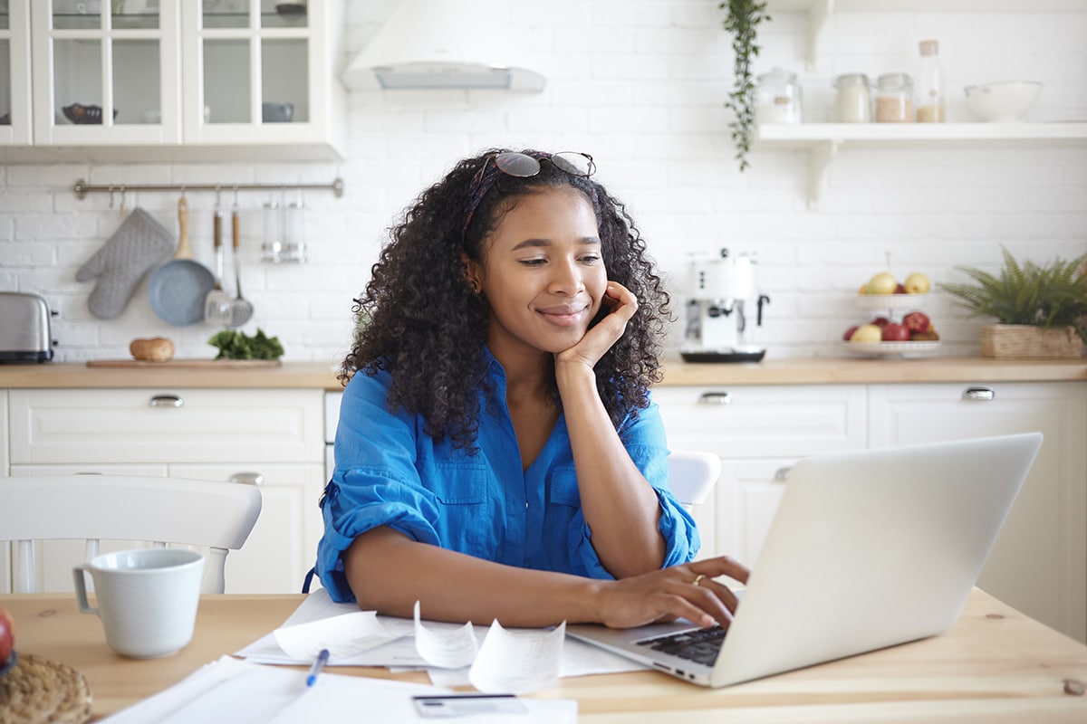 Woman sitting at a kitchen table smiling while typing on her laptop