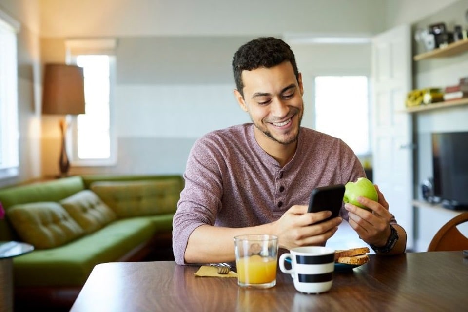 A man smiles while holding an apple in one hand and looking at his phone, embodying a healthy lifestyle and financial freedom.