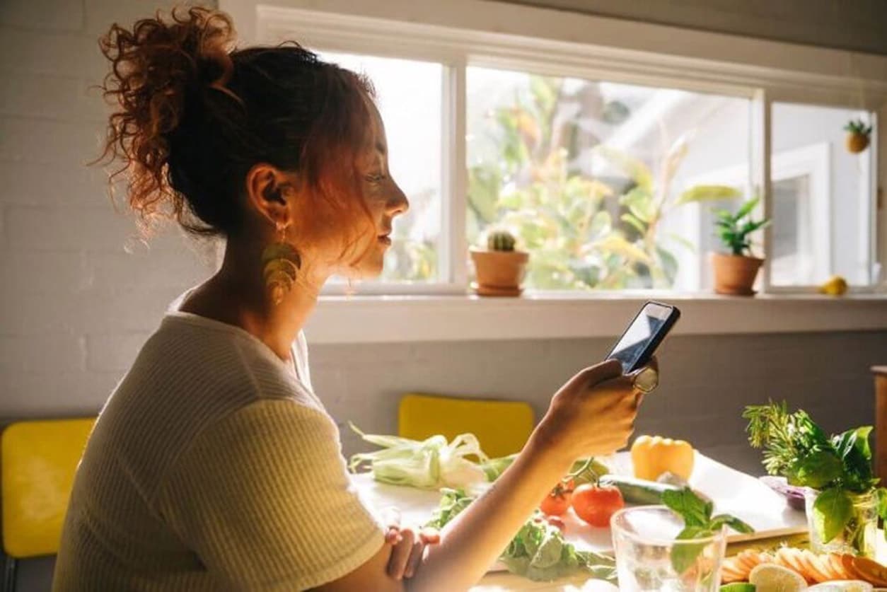 A woman, seated in front of a window, examines her phone, reflecting on the credibility of debt relief programs.