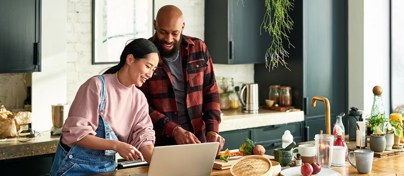 Woman and man are in the kitchen hanging out, smiling and looking at a laptop