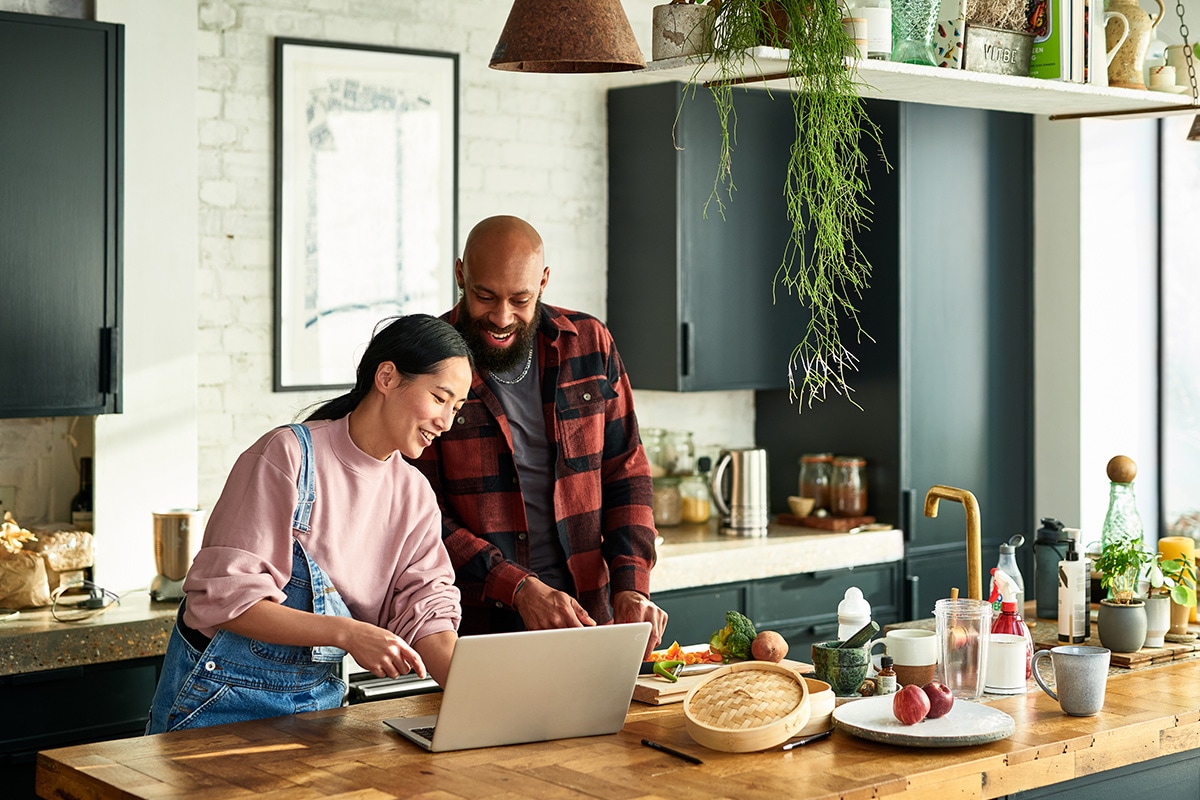 Woman and man are in the kitchen hanging out, smiling and looking at a laptop