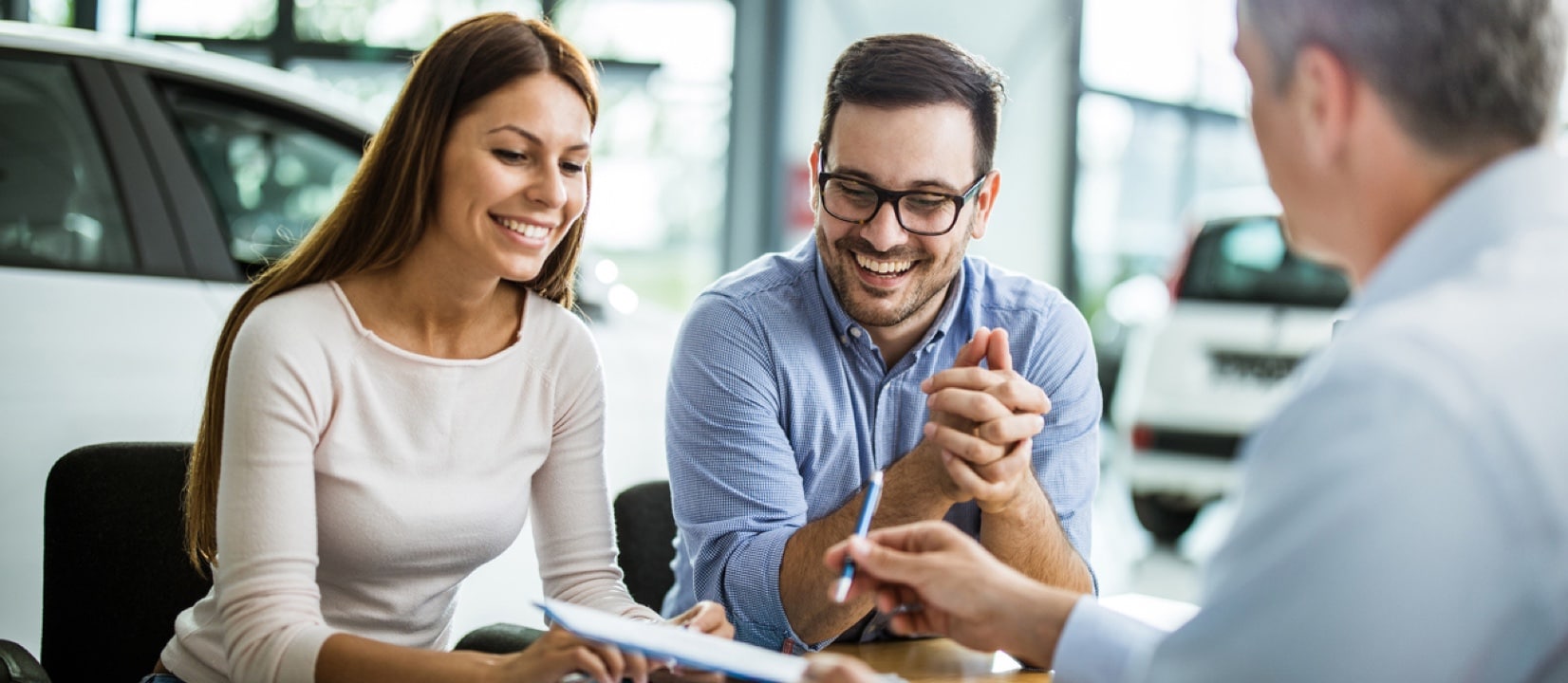 Couple is at the dealership is talking to a salesman and looking through paperwork