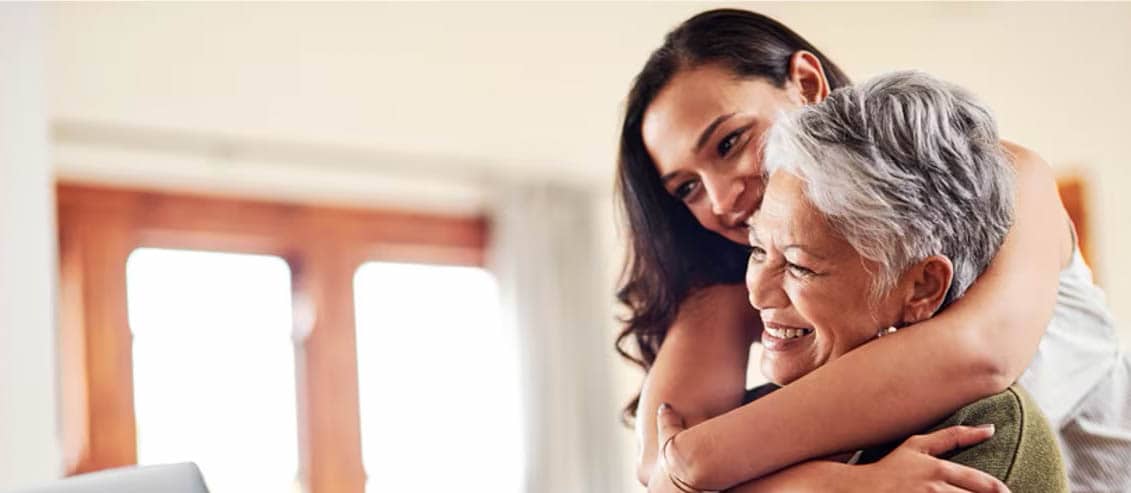 A woman embraces an older woman while seated in front of a laptop, conveying warmth and connection between generations.