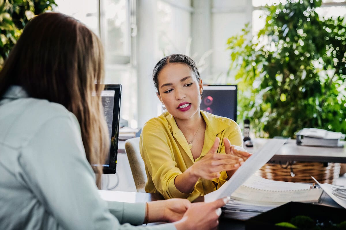 Two women are talking in the office and looking through paperwork.