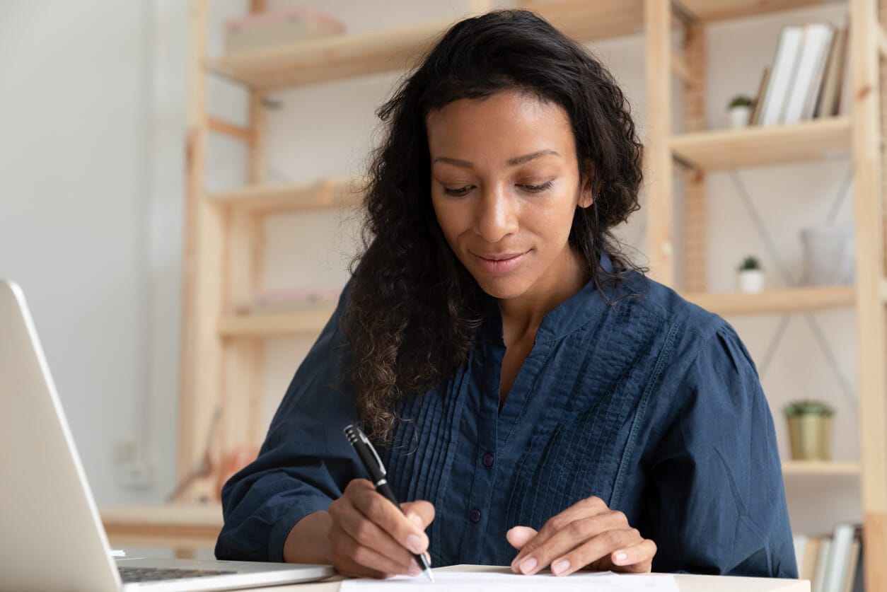 A woman sits at her desk writing in her notebook with her laptop open next to her