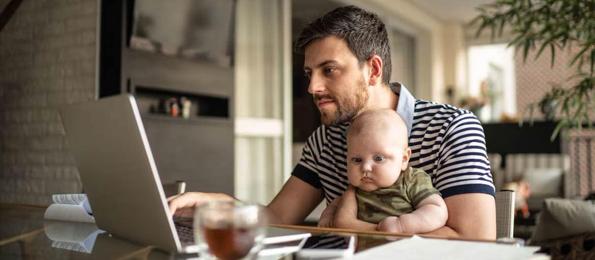 A man and a baby sit at a table, engaged in financial research on a laptop, showcasing a moment of learning and bonding.