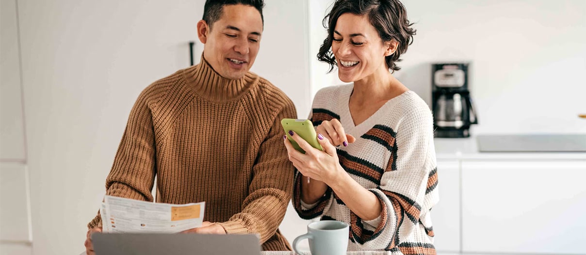 Couple sitting at a kitchen island, looking at a phone with papers and a laptop in front of them