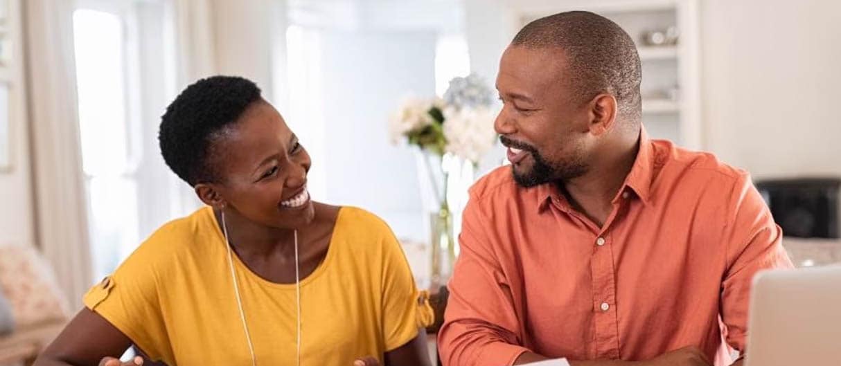 smiling couple sitting at a wooden table with a notebook, papers, and a laptop