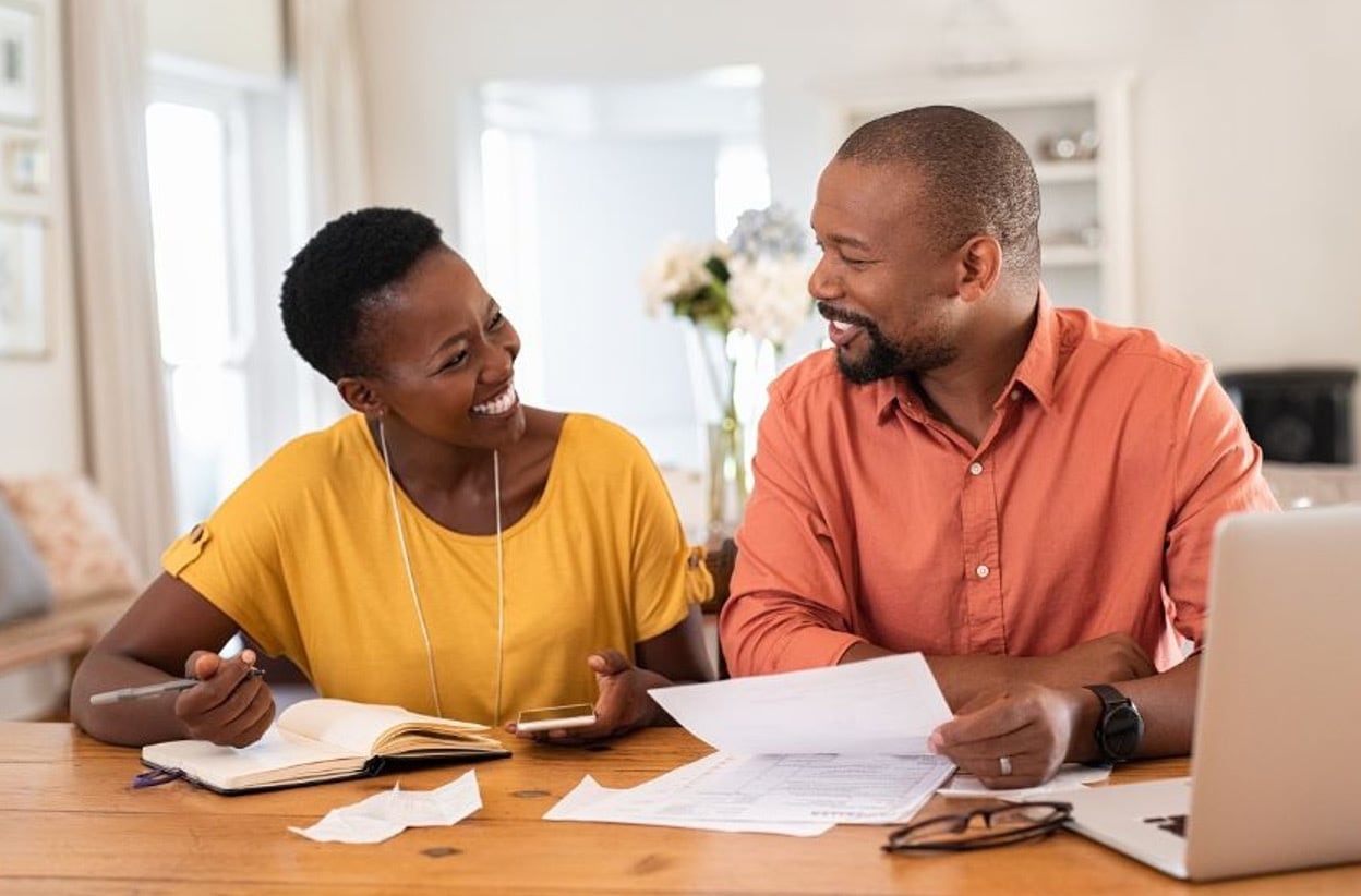 smiling couple sitting at a wooden table with a notebook, papers, and a laptop