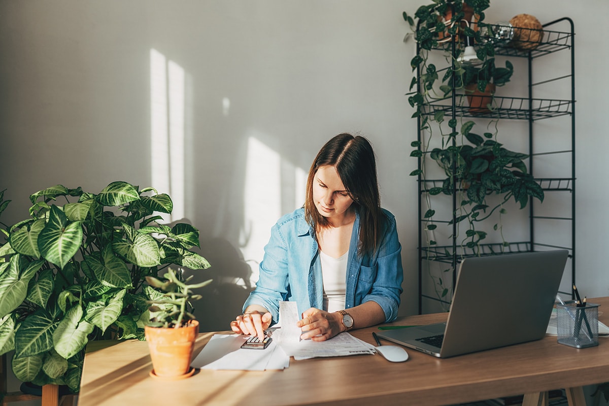 A woman sits at a desk with a laptop and notebook, contemplating fixed and variable expenses for financial planning.