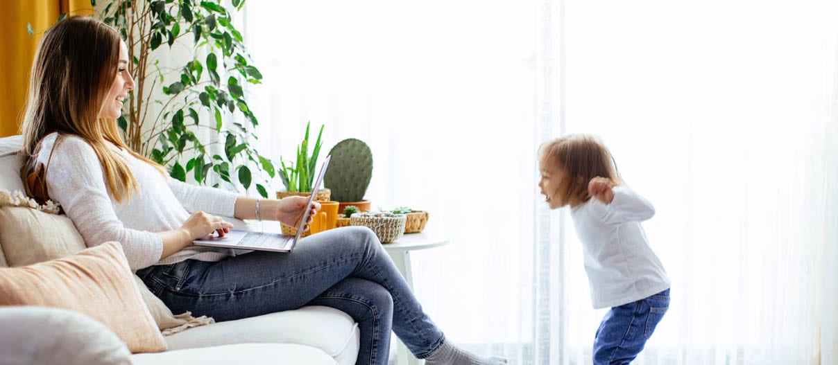 Woman sitting on a couch with laptop open smiling at a child jumping in front of her