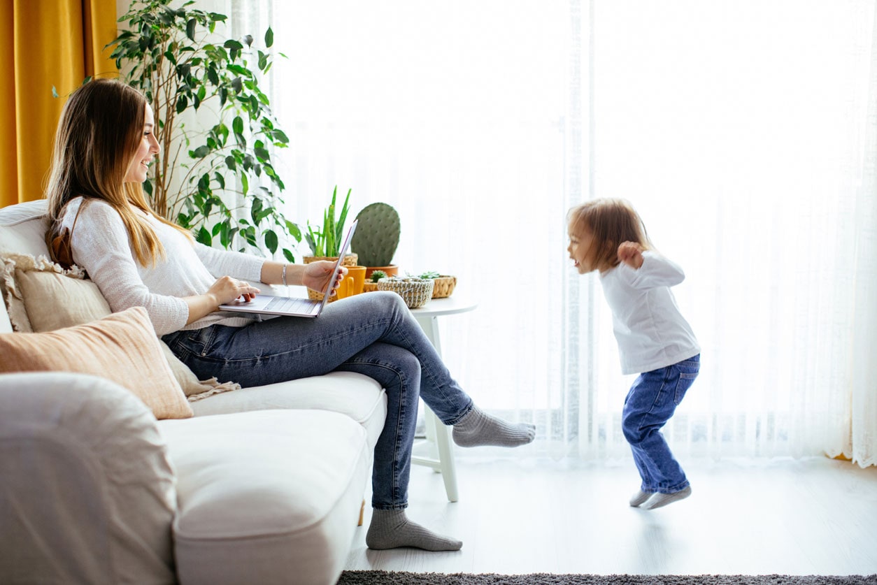Woman sitting on a couch with laptop open smiling at a child jumping in front of her