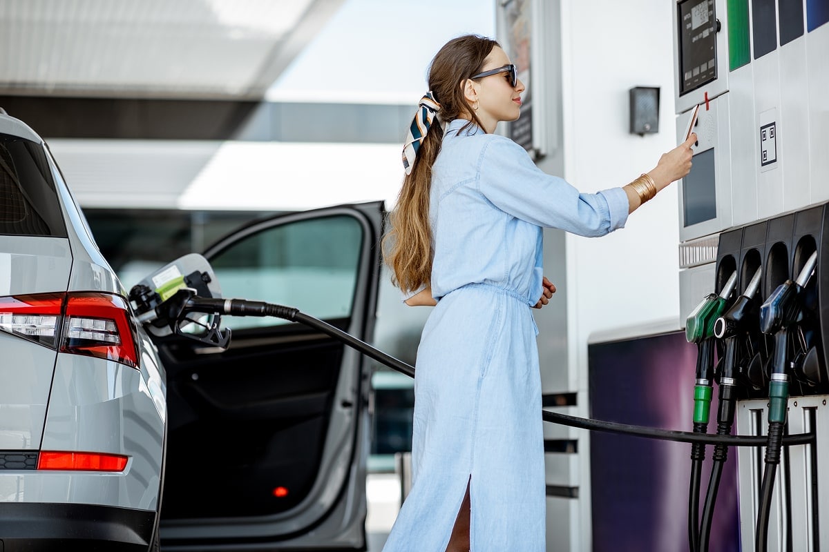  A bright, daytime lifestyle photo of a driver standing next to their car at a gas station. They are focused on the payment screen at the pump, holding a credit card in one hand and their wallet in the other.