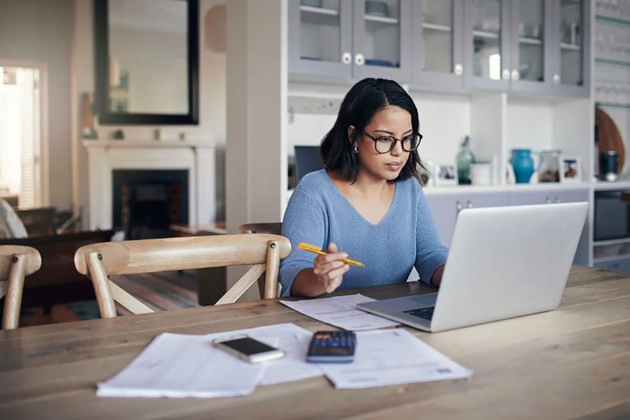 A woman in glasses sits at a table, focused on her laptop and papers, exploring strategies to reduce debt.