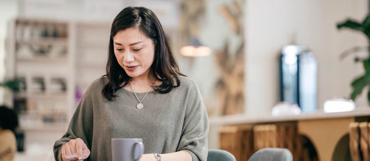A woman sits at a cozy table with a coffee mug, using a tablet.