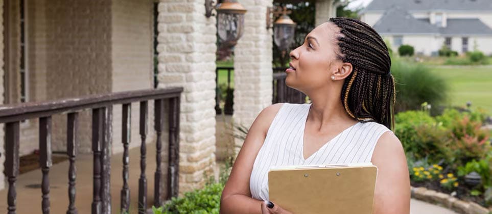A woman standing in front of a house, holding some documents, and looking at the home.