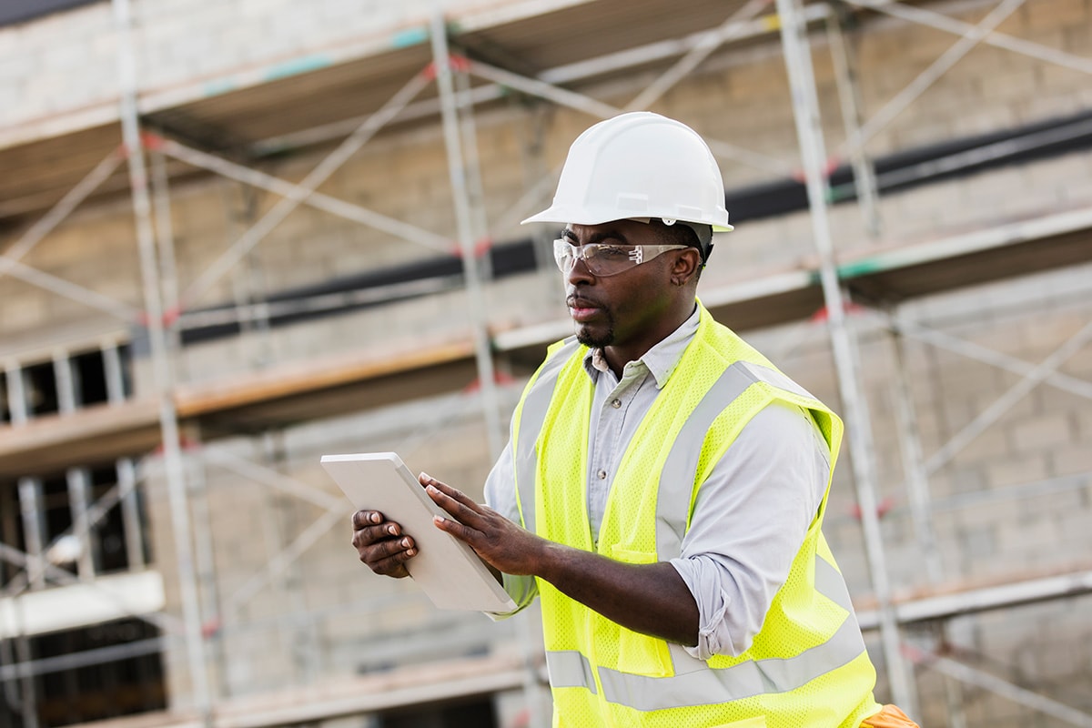 A man in a hard hat and safety vest examines a tablet, likely assessing home inspection costs and details.