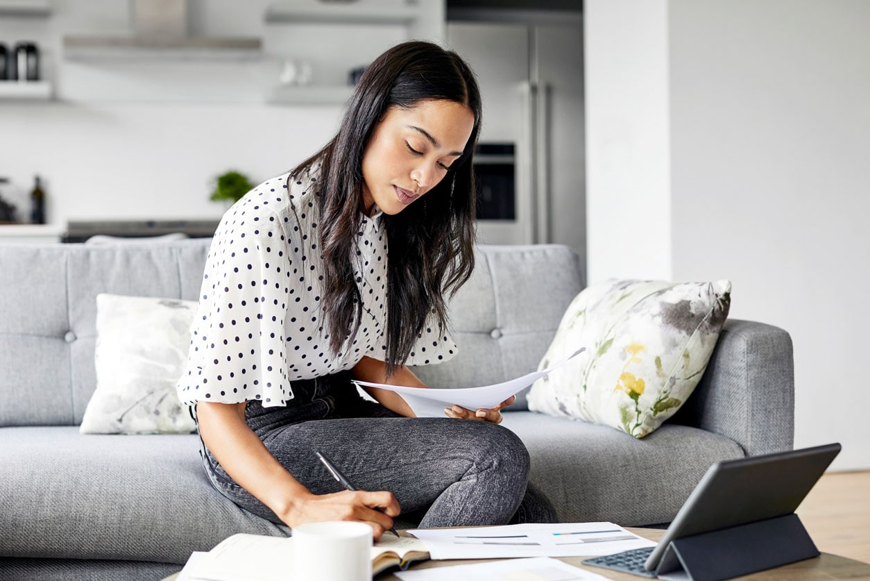 A woman sitting on the edge of her living room couch doing paperwork