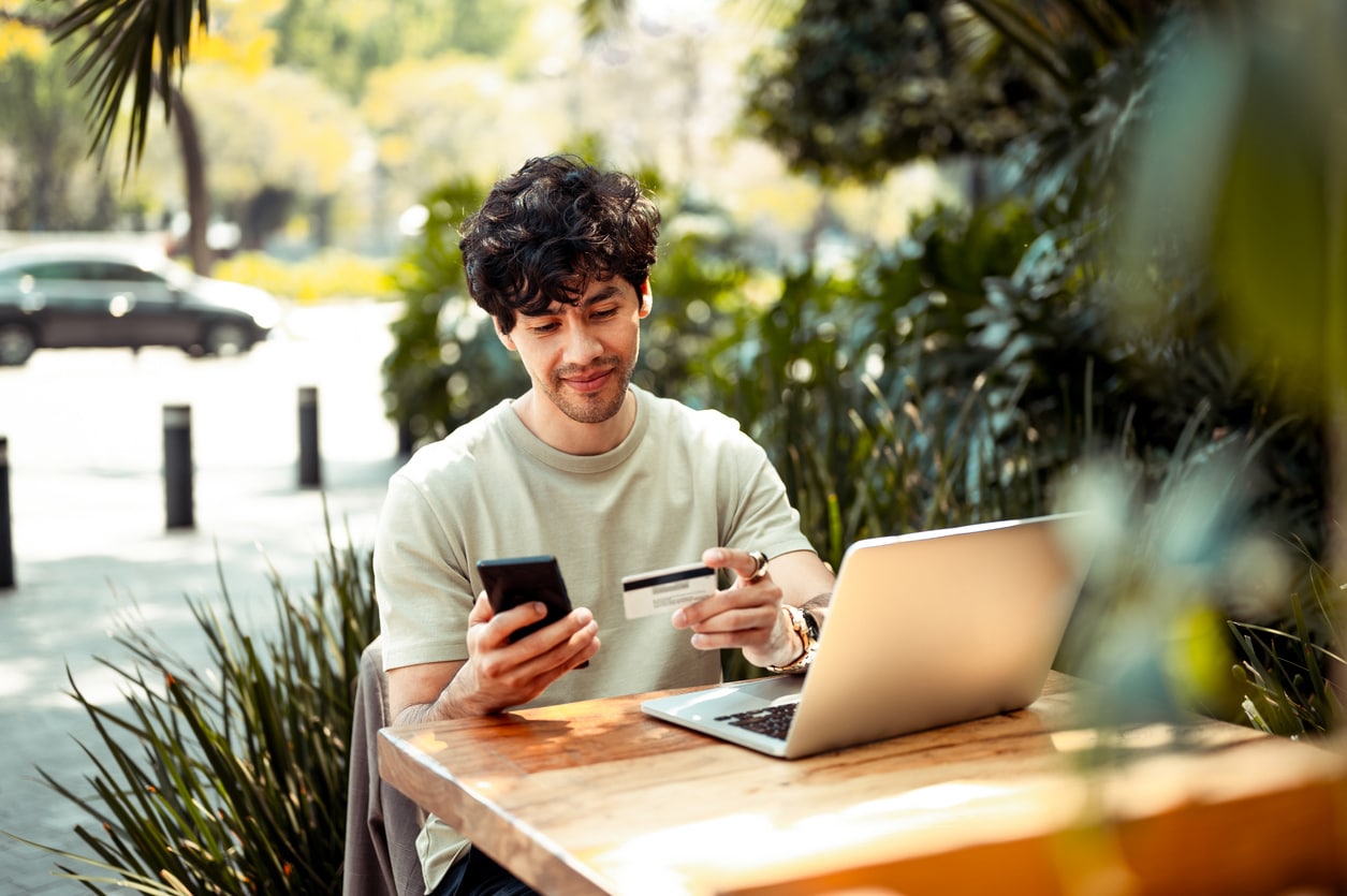 A man seated at a table, using his phone and a credit card, possibly researching how to dispute his credit report.