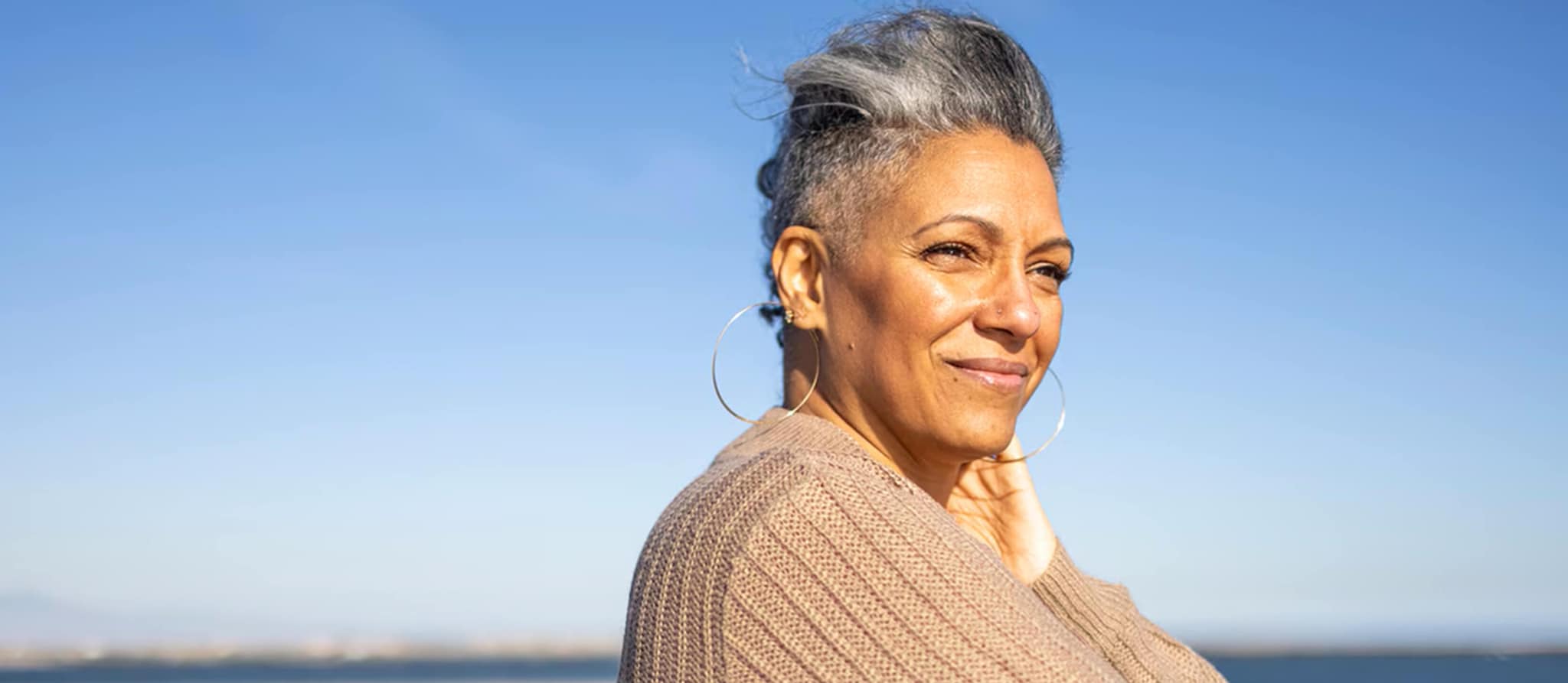 An older woman with gray hair stands by the ocean, reflecting on life and financial security as she approaches age 60.