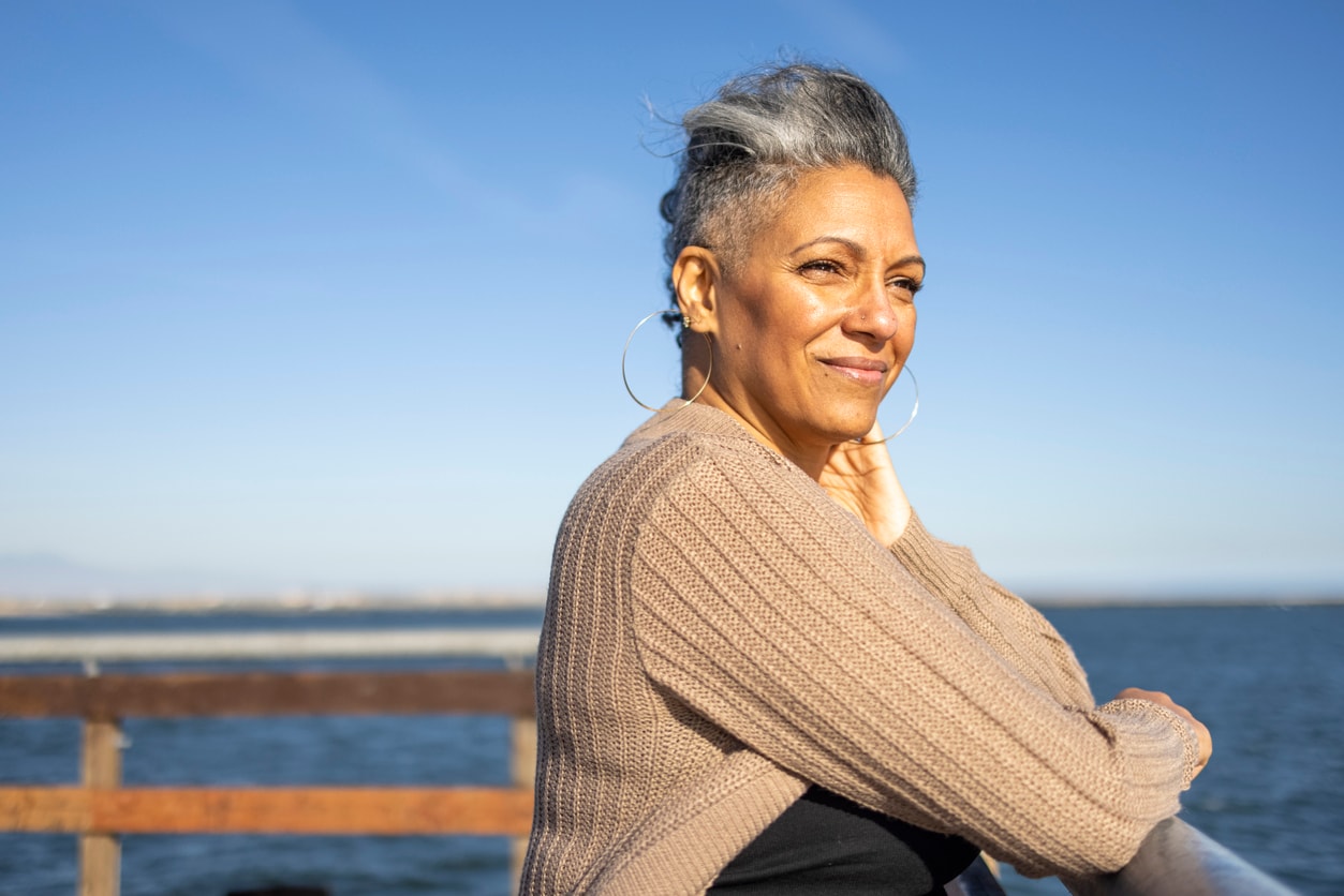 An older woman with gray hair stands by the ocean, reflecting on life and financial security as she approaches age 60.
