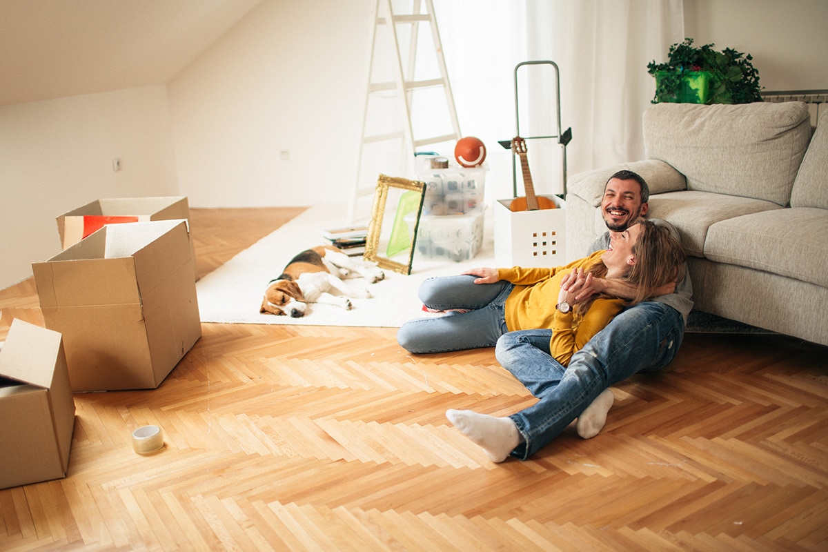 A man and woman sit on the living room floor surrounded by boxes, contemplating their budget for rent and living expenses.