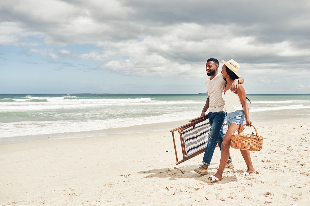 A couple strolls along the beach, carrying a picnic basket, embodying leisure and financial freedom through mindful living.