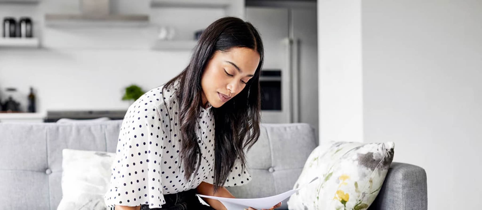 A woman sits on a couch, focused on reading a document about balancing her checkbook effectively.