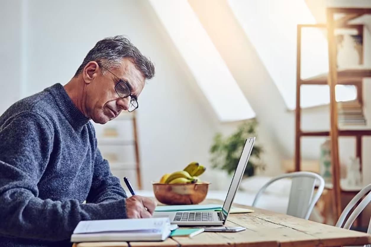 A man writes in a pad that is next to his laptop while sitting at his desk