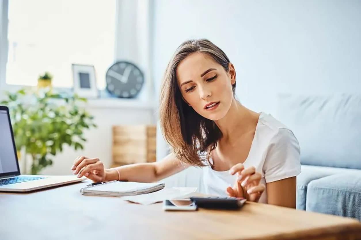 Woman sitting at her coffee table calculates her debt using her laptop, notepad, and calculator.